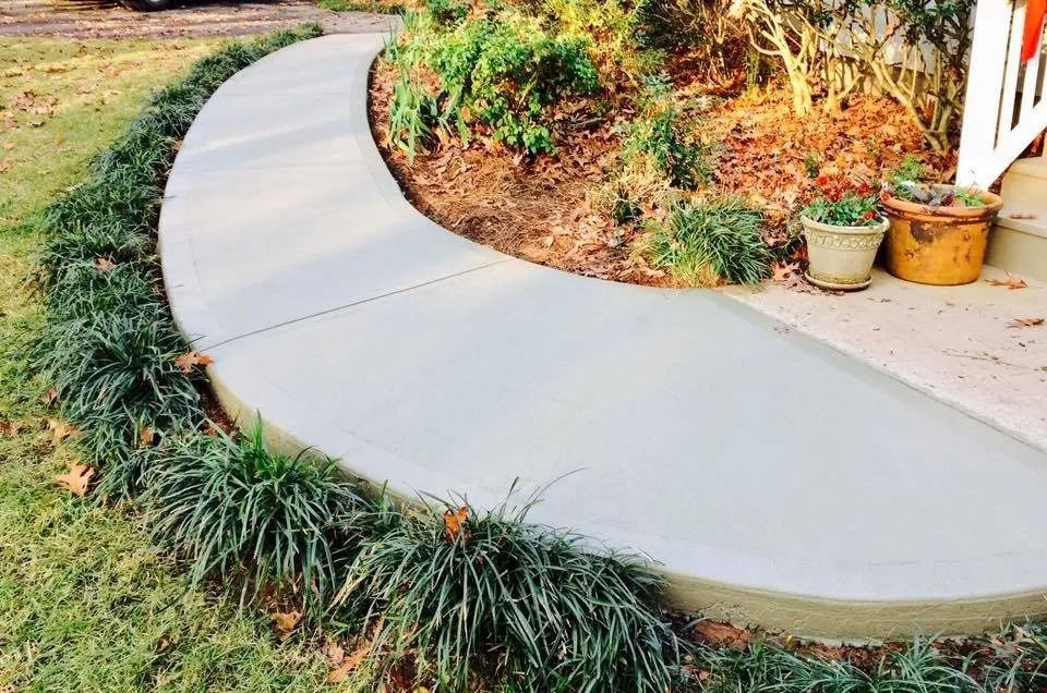 A curved concrete walkway bordered by green ornamental grass, with potted plants on a porch in the background.
