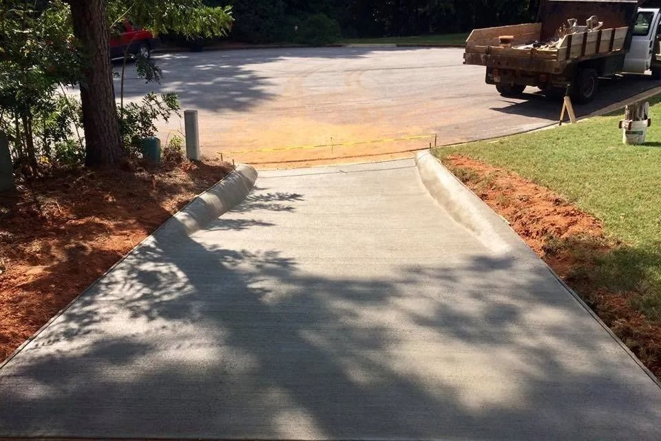 A newly poured concrete driveway ramp slopes downward toward a paved lot with a dump truck parked in the background.