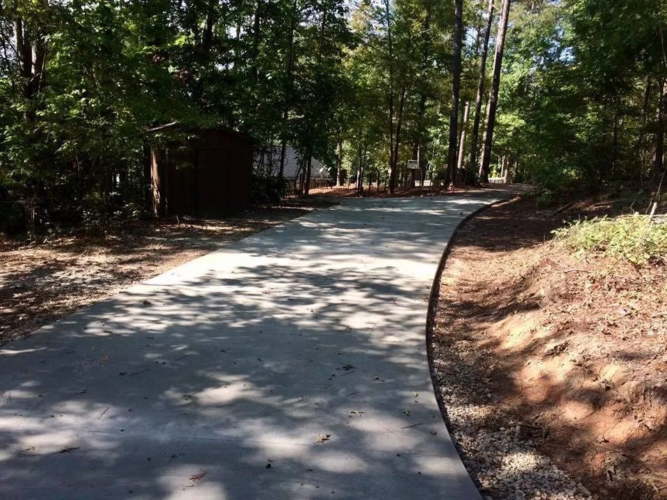 A curved concrete driveway winds through a wooded area with trees casting shadows on the surface.