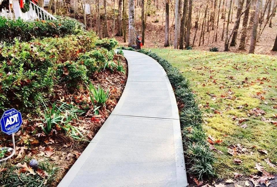 A light gray concrete walkway curves through a yard with green bushes on the left and a grass lawn on the right.