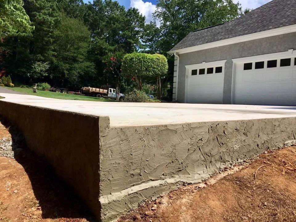 A concrete driveway extension with a freshly parged side wall, set in front of a garage surrounded by trees and dirt.
