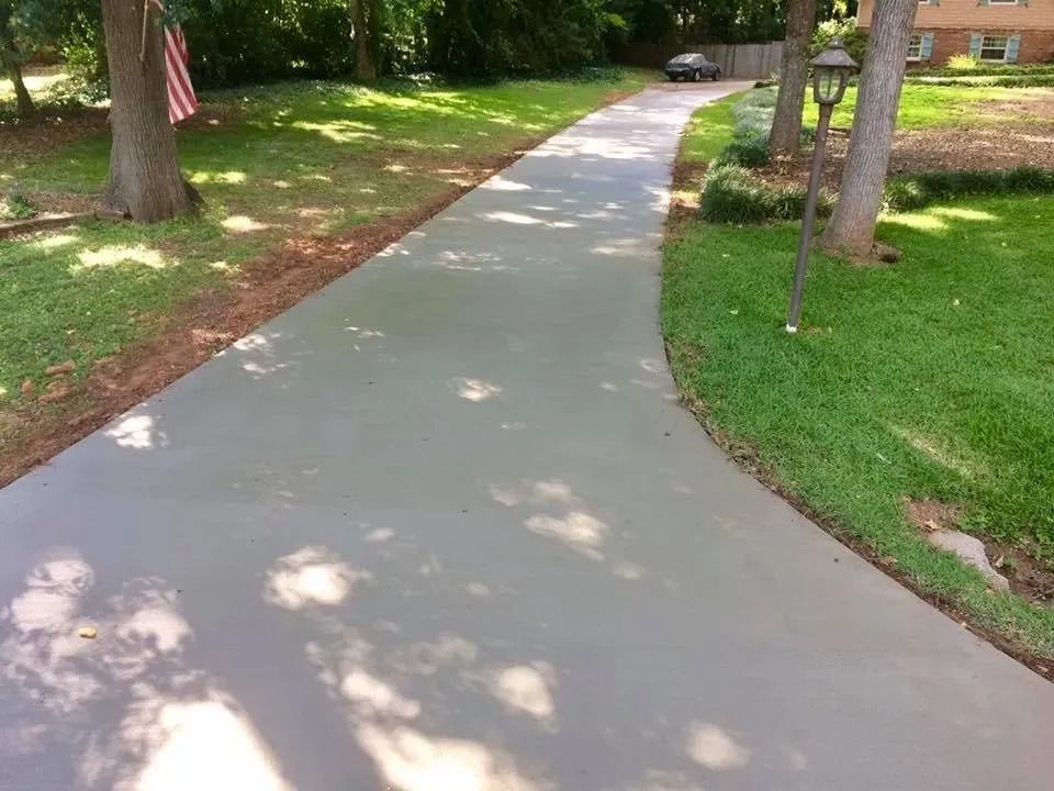 A smooth, gray concrete driveway curves toward a house, flanked by green lawns and trees on a sunny day.