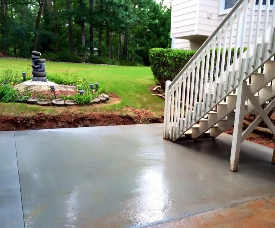 A freshly poured concrete patio adjacent to wooden stairs and a grassy yard with a tiered stone feature.