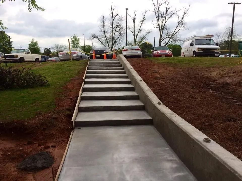 A set of newly poured concrete stairs with retaining walls leading up a grassy hill to a parking lot.