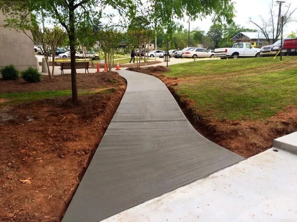 A newly poured concrete walkway curves through a grassy park area near a parking lot.
