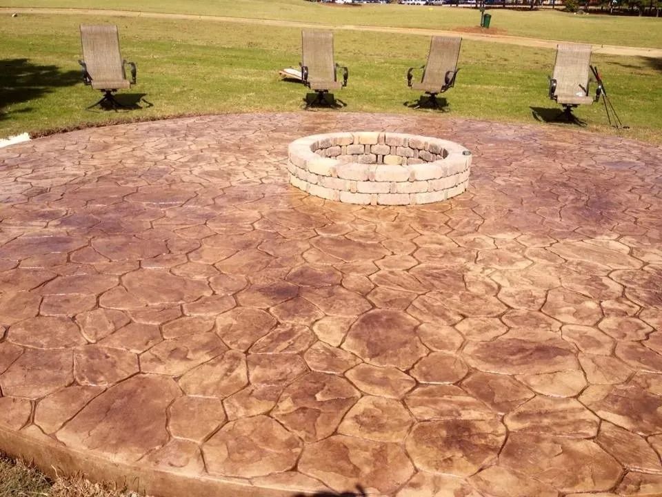 A circular stamped concrete patio with a stone fire pit in the center, featuring four chairs on the surrounding grass.