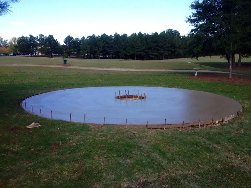 A newly poured circular concrete pad with exposed rebar in its center, situated on a grassy park lawn.