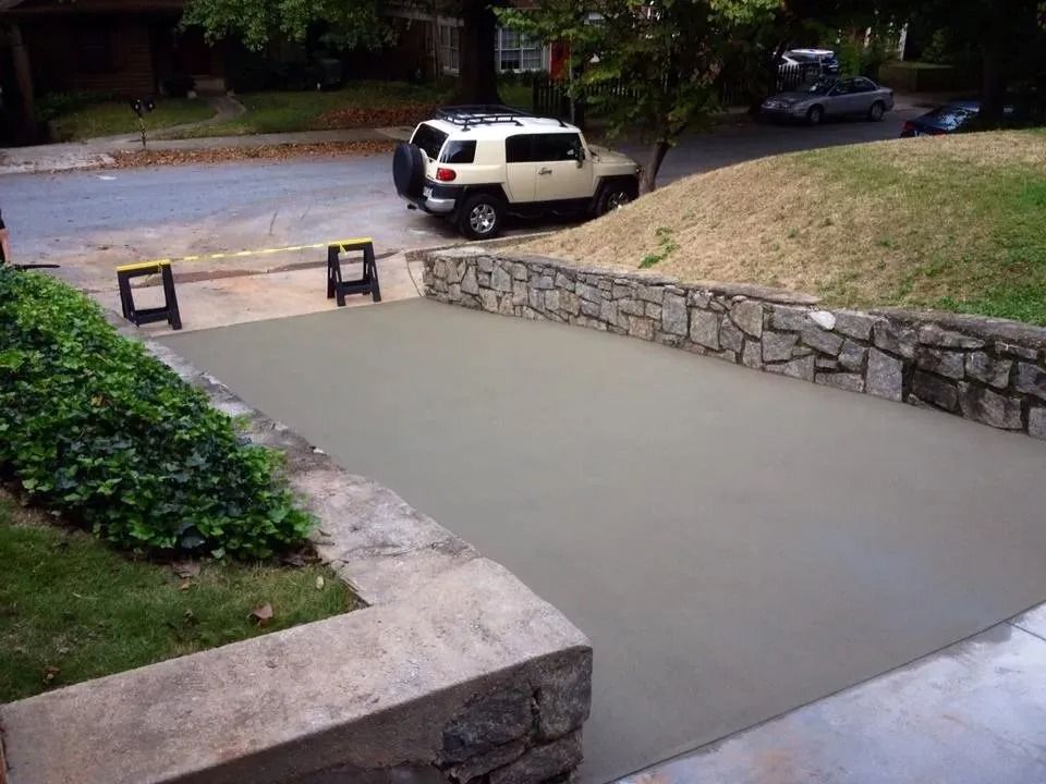 A freshly poured, smooth concrete driveway framed by stone retaining walls, with two yellow sawhorses in the background.
