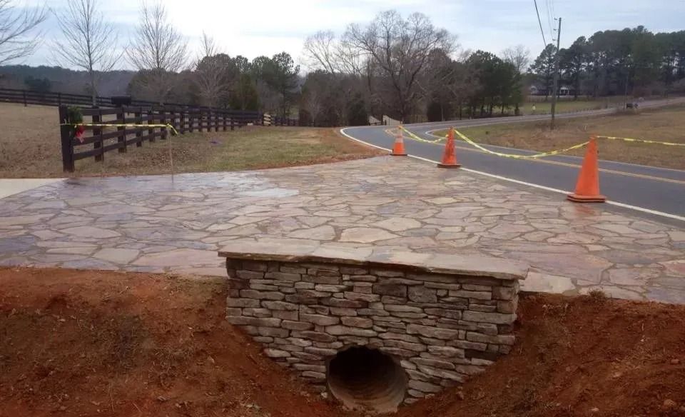 A stone-faced culvert entrance in a red-dirt ditch next to a patterned concrete driveway and a paved rural road.