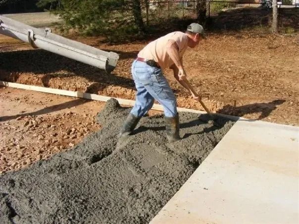 A person uses a tool to spread wet concrete from a chute into a prepared area outdoors.