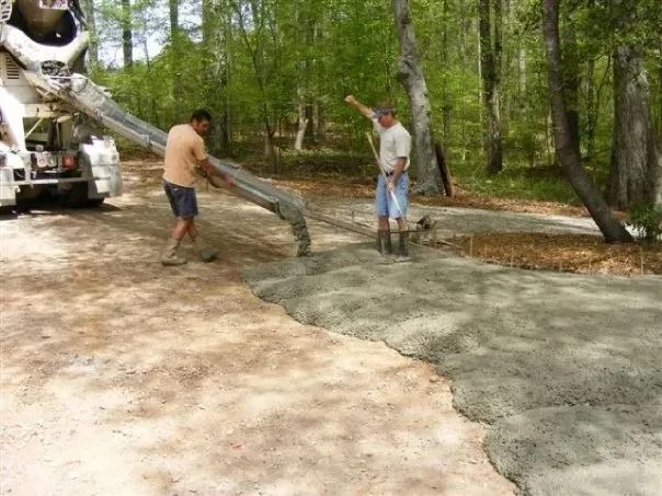 Two people use a concrete truck chute to pour wet cement onto a wooded construction site.