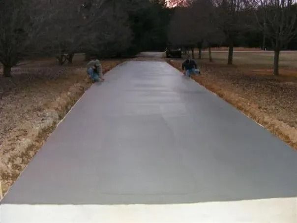 Two people finish smoothing a freshly poured, gray concrete driveway lined with trees in a wooded area.
