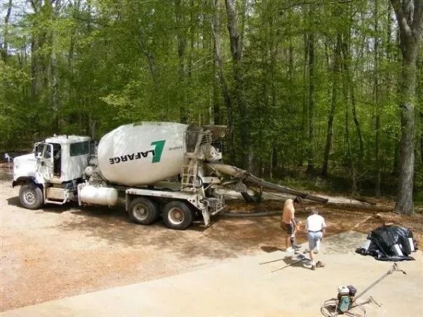 A Lafarge cement mixer truck is pouring concrete at a wooded construction site with two workers nearby.