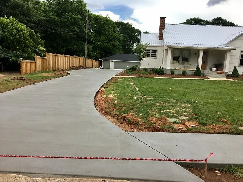 A newly poured concrete driveway leads to a white house and detached garage, next to a wooden fence and grassy front yard.