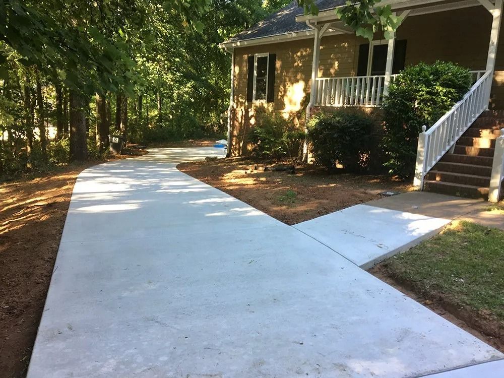 A newly poured concrete driveway leads to the front porch steps of a house surrounded by trees.