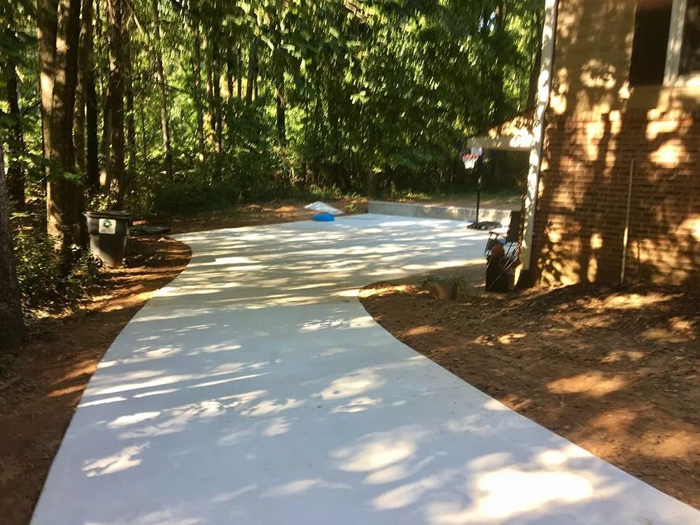 A newly poured concrete driveway leads to a basketball hoop beside a brick house near a wooded area.