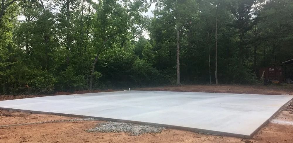 A large, rectangular, light-gray concrete slab sits in a clearing surrounded by green trees on a dirt lot.