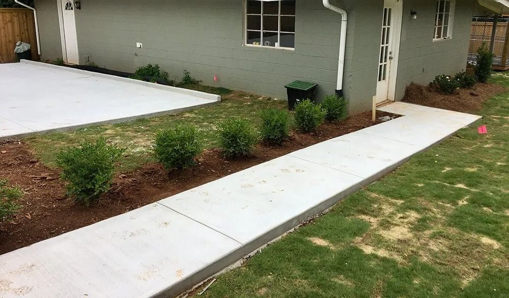 A gray house exterior with a new concrete sidewalk leading to a doorway, lined with small green shrubs and mulch.