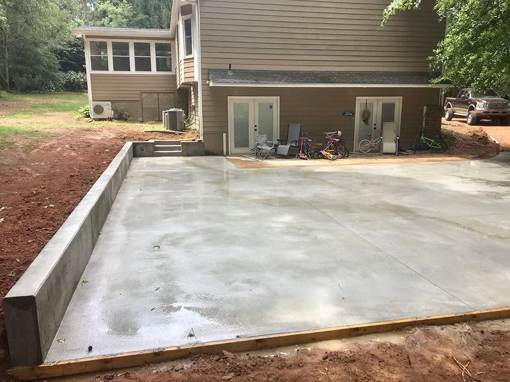 A newly poured concrete patio with a retaining wall and wooden formwork against the brown exterior of a two-story house.
