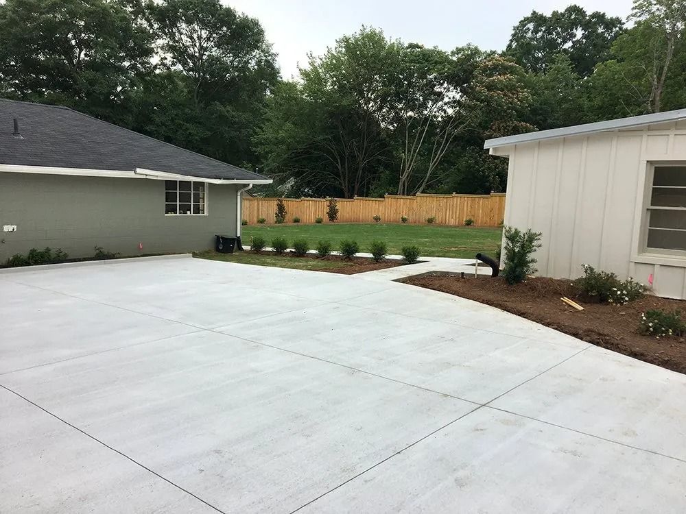 A newly poured concrete driveway situated between a grey house and a white building, leading toward a wooden fence.