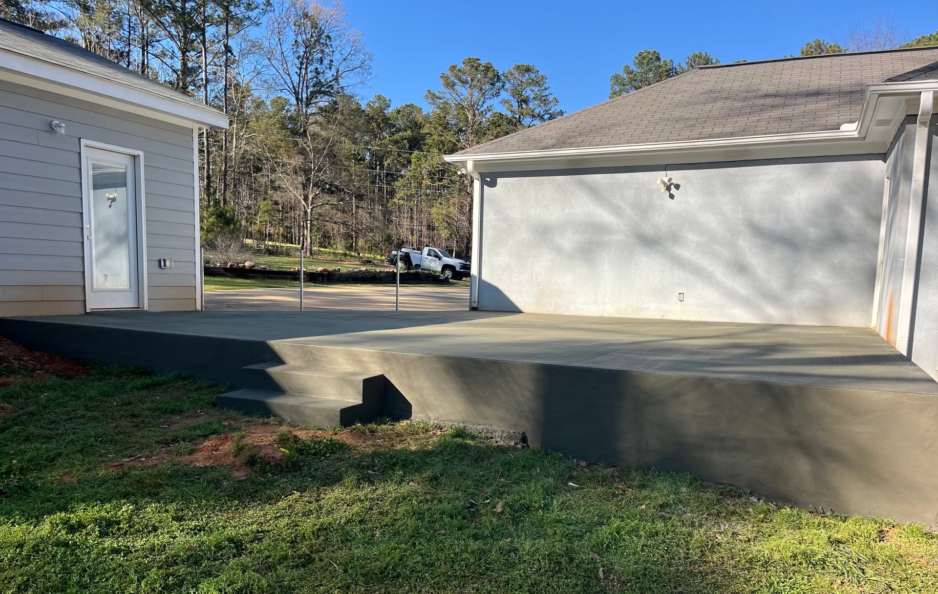 A new concrete patio with stairs sits between two sections of a light-colored house in a yard with trees in the background.