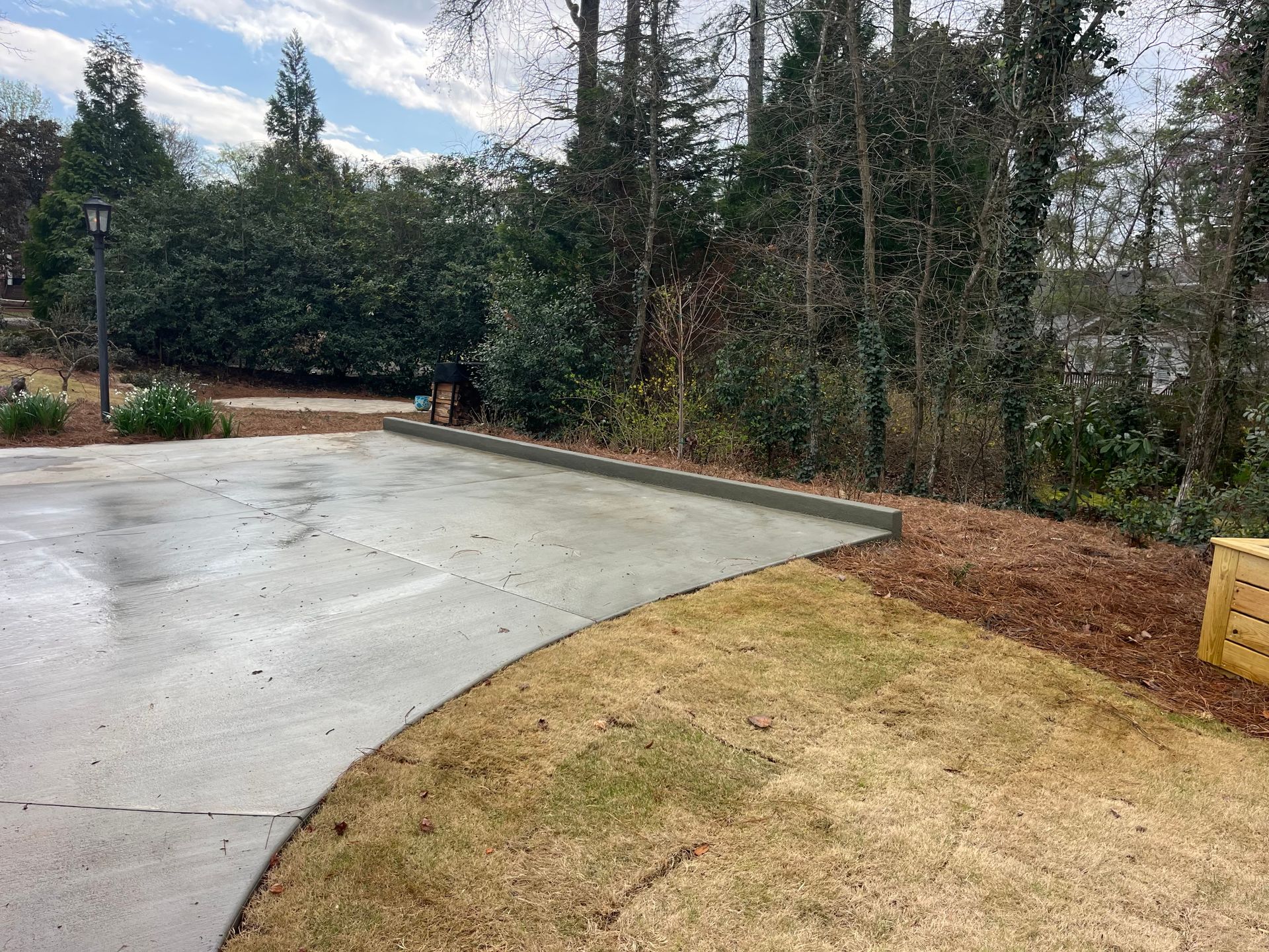 A concrete driveway edge meets a mulched garden bed and a patch of dormant grass under a bright, partly cloudy sky.
