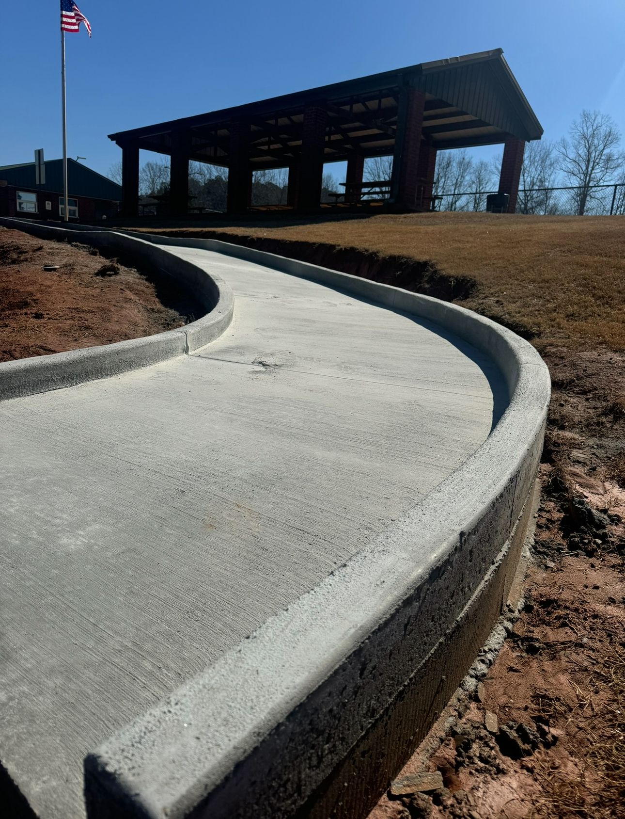 A concrete sidewalk with a curved curb leading toward a park pavilion under a clear blue sky.