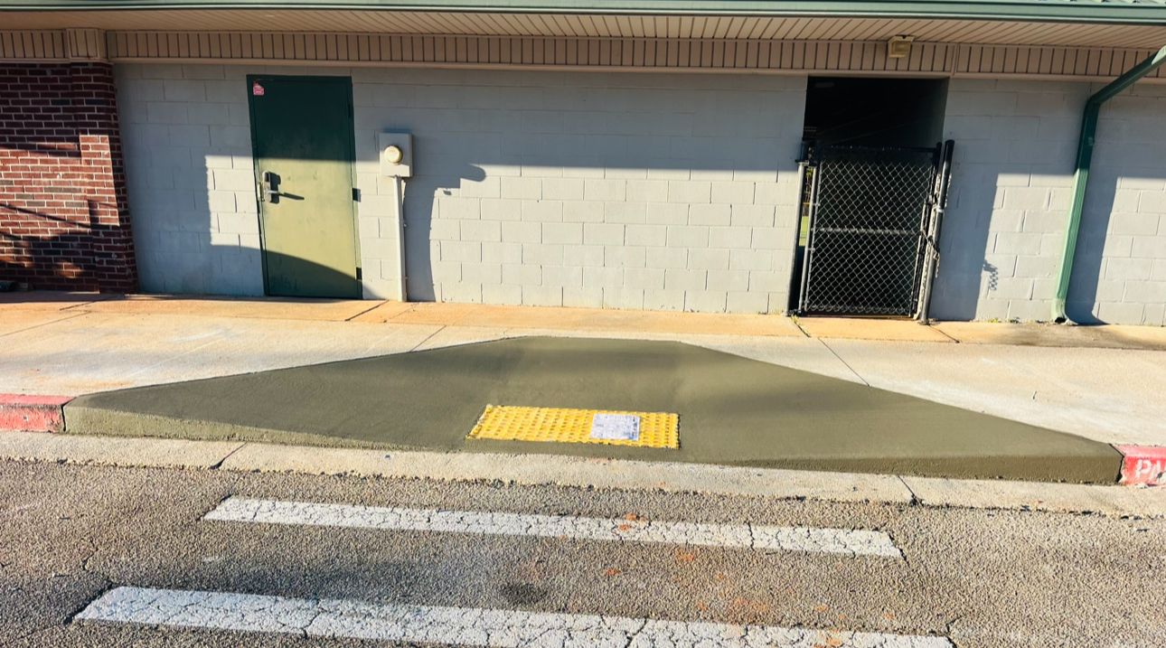 A concrete curb ramp with a yellow tactile warning strip leading to a building entrance with two doors.