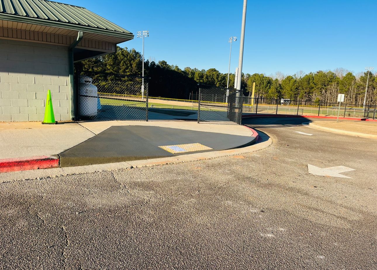 A freshly paved concrete walkway leads to a building entrance next to a gravel parking lot with painted directional arrows.