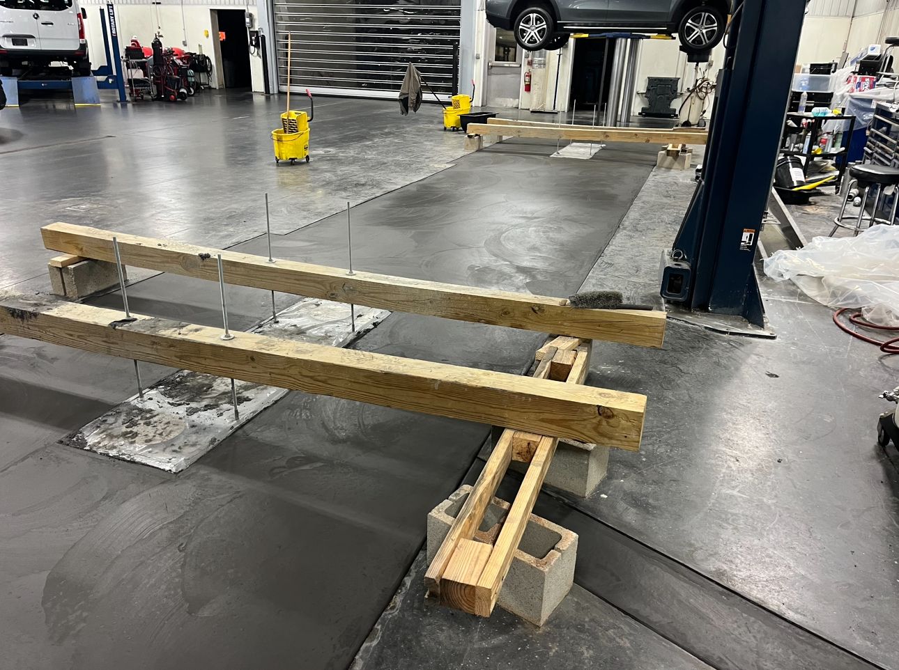Wooden beams suspended over a concrete floor in an auto repair shop, supported by cinder blocks and threaded rods.