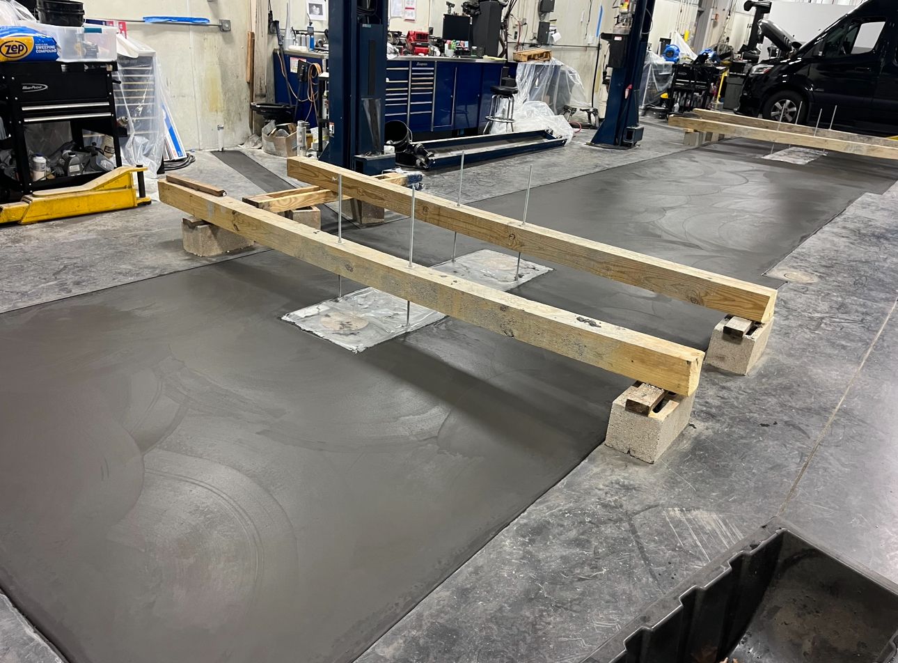 Two wooden beams supported by concrete blocks over a freshly poured concrete patch in a workshop setting.