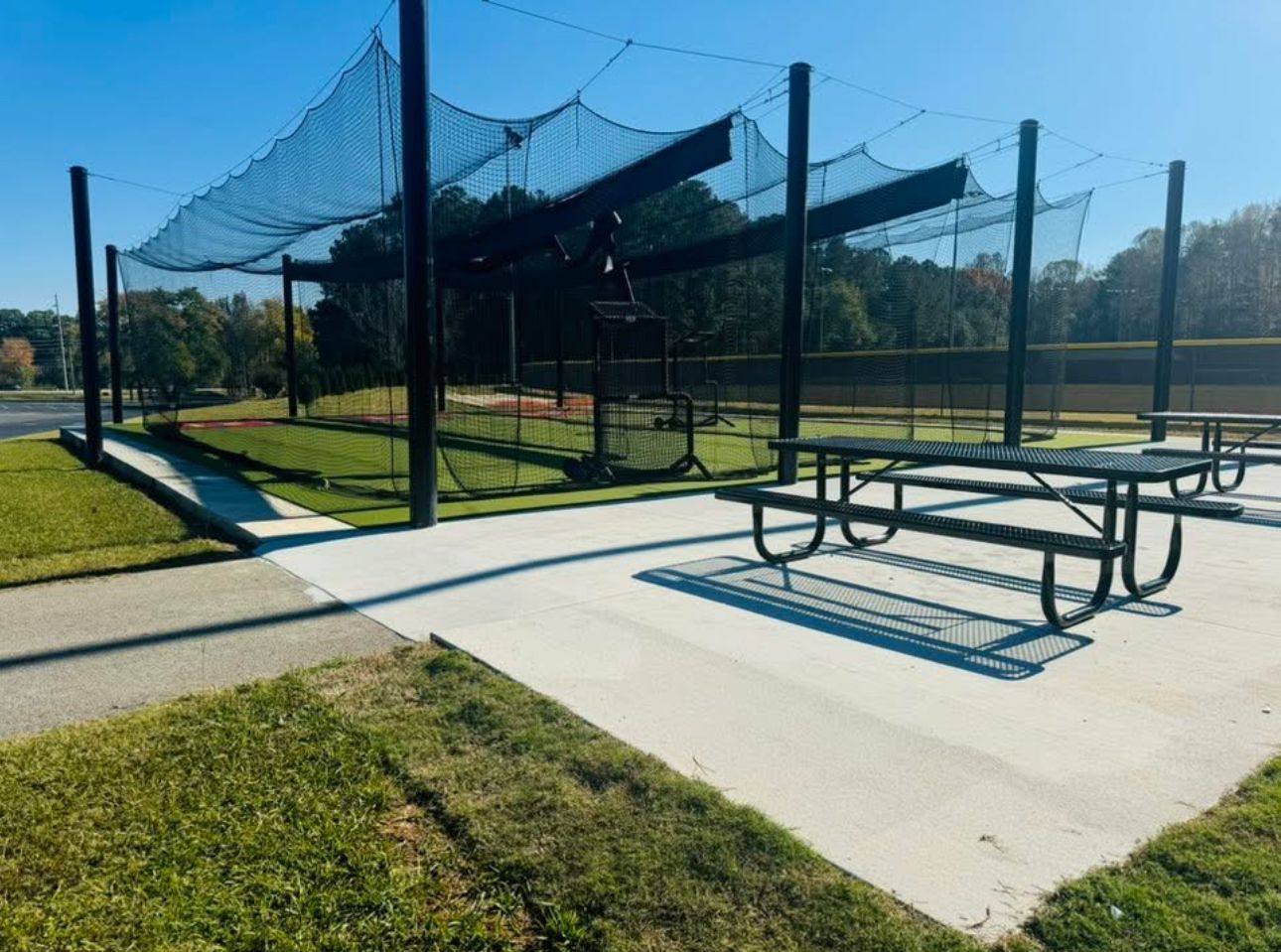 A black-netted batting cage structure next to a picnic table on a concrete pad at a park under a clear blue sky.