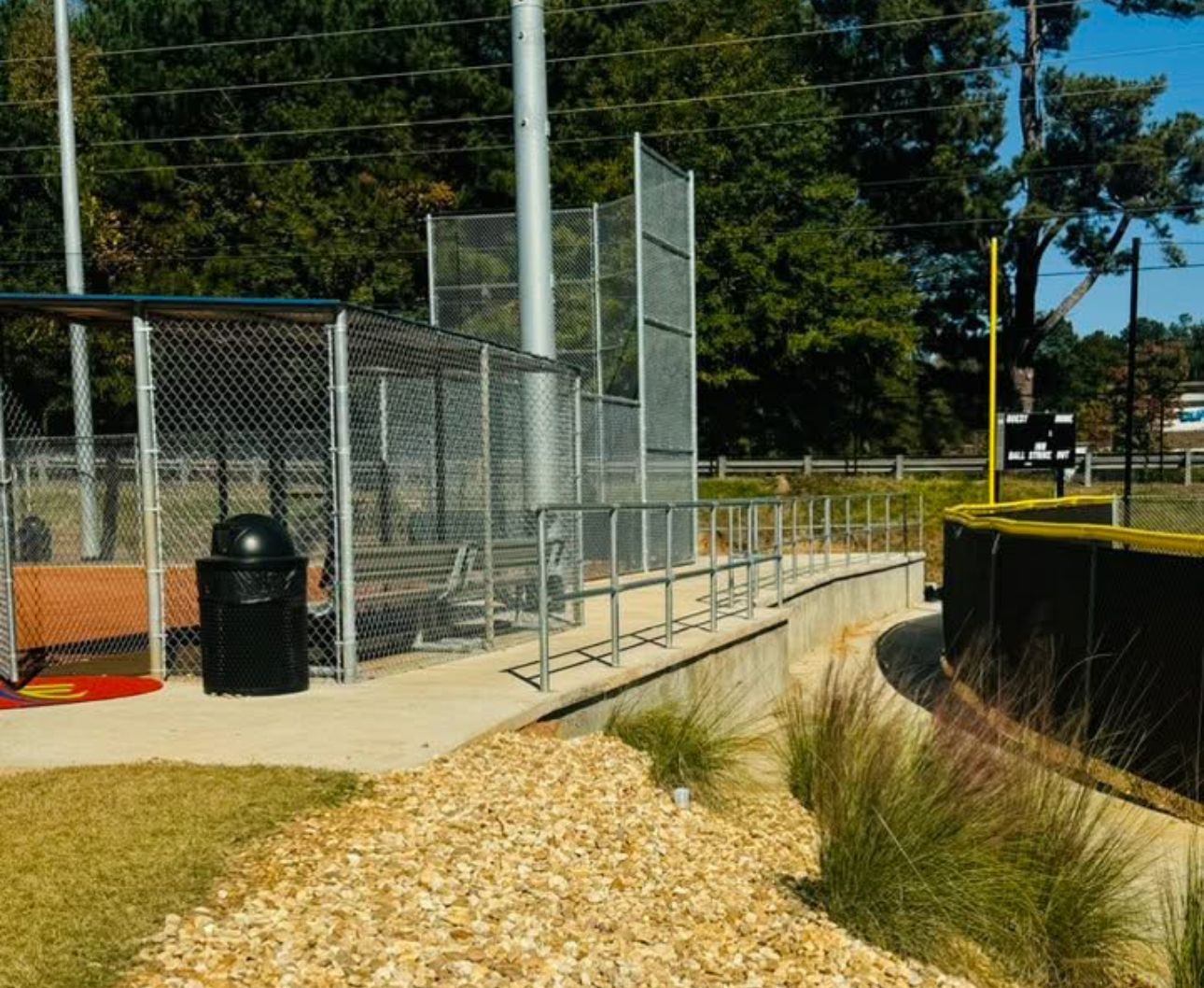 A fenced baseball dugout with a black trash bin, a concrete walkway, and landscaped gravel area under a bright blue sky.