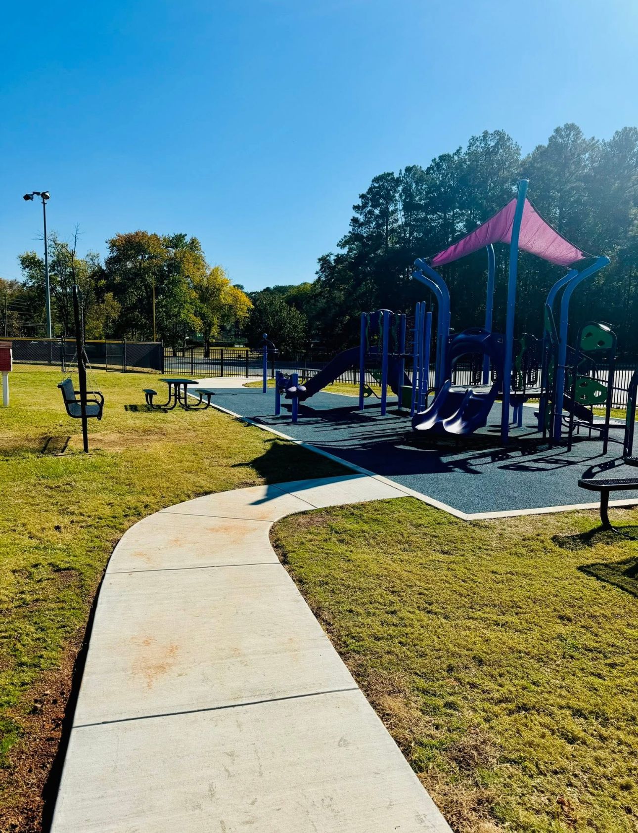 A concrete path leads to a blue and pink playground structure on a rubber surface in a sunny, grassy park.