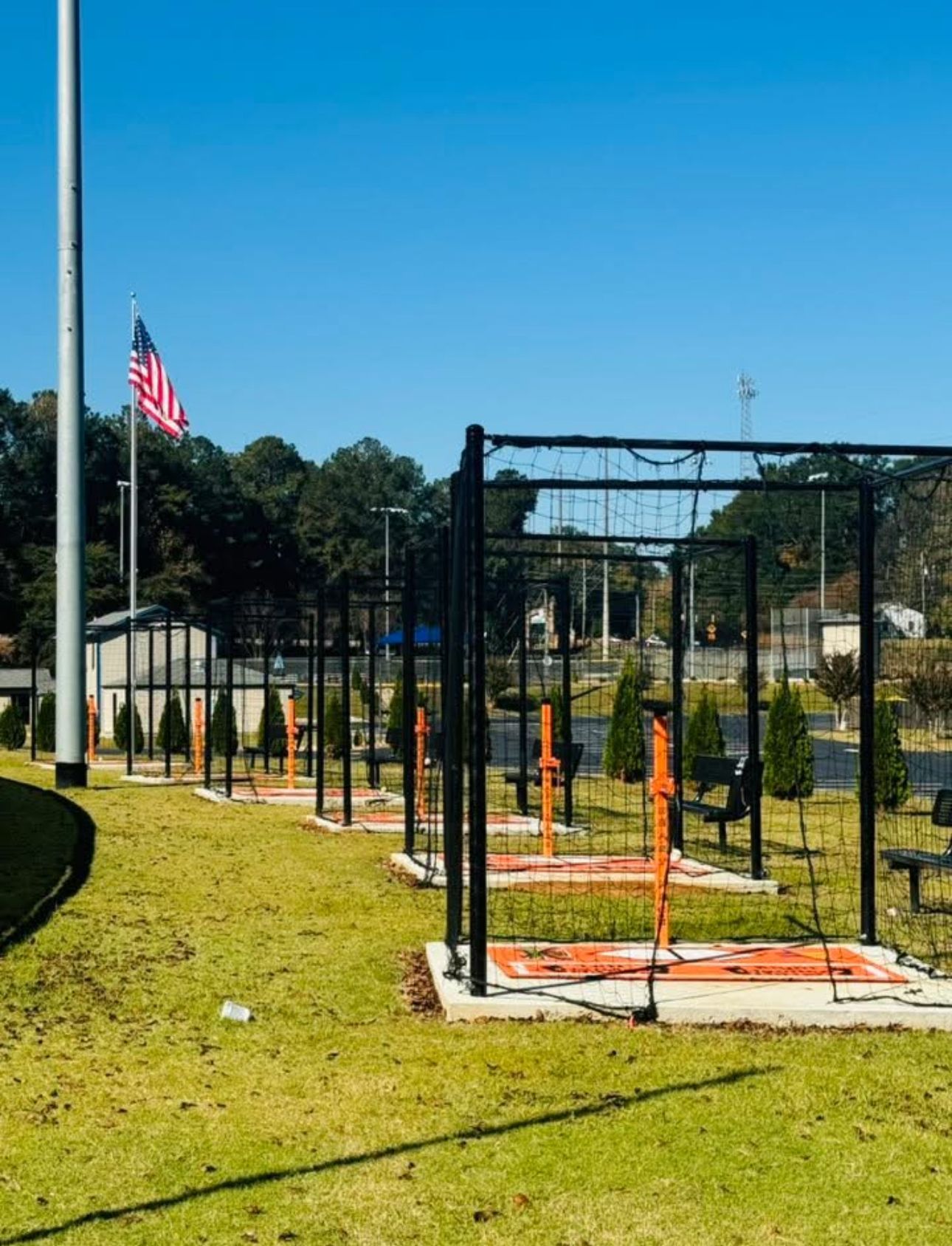 Outdoor fitness course with black metal frames and orange equipment on a grassy field under a clear blue sky with a flag.