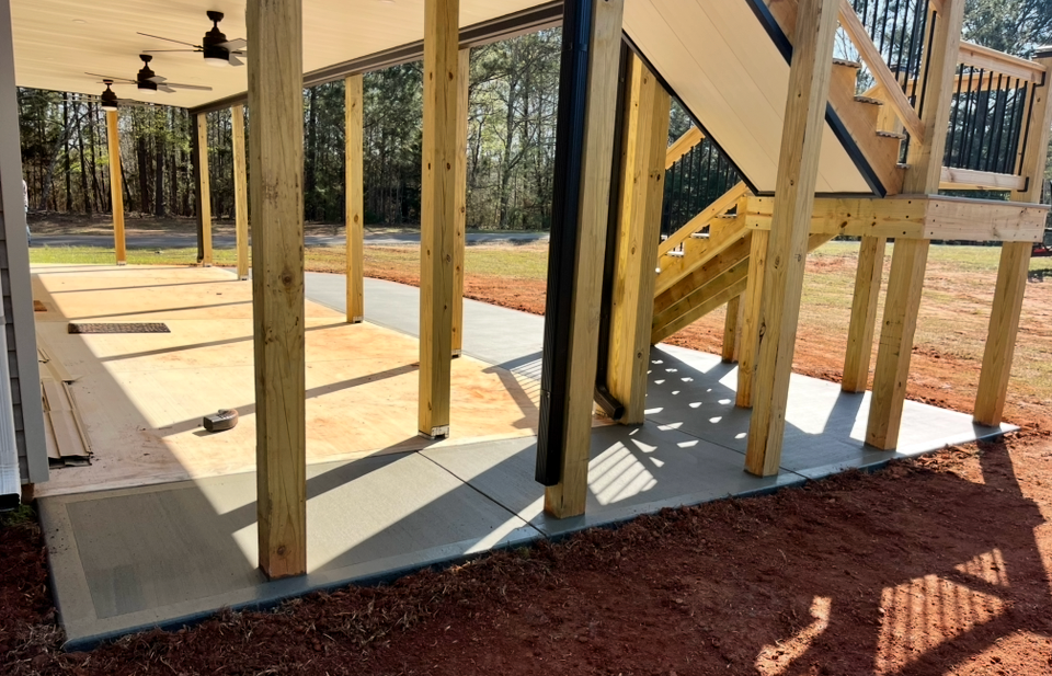 New deck structure with stairs and concrete patio flooring supported by vertical wooden posts in a backyard.