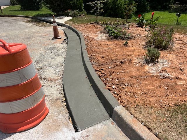 Newly poured concrete curb along a dirt landscaping area, with orange traffic cones visible in the foreground.