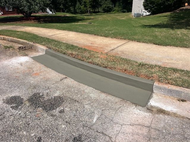 A fresh concrete curb ramp installed at the edge of an asphalt street, adjacent to a sidewalk and grassy lawn.