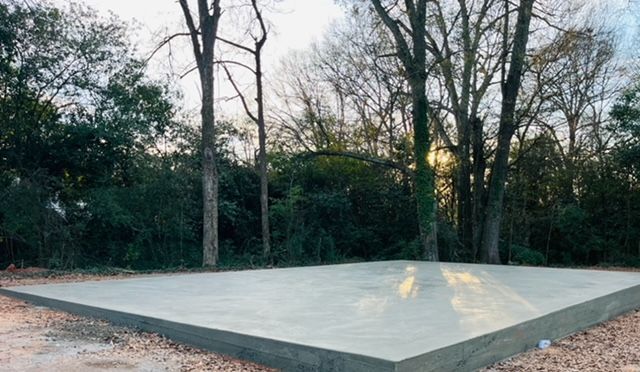 A rectangular concrete slab foundation set in a wooded area with trees in the background under a soft, golden light.