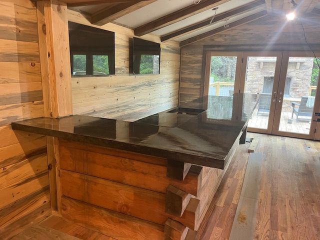A rustic bar with a dark, polished countertop and log-style base, set in a room with wood paneling and a patio view.