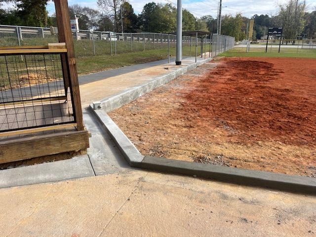 A concrete curb edge separates a paved walkway from a reddish-brown dirt baseball field at a park.