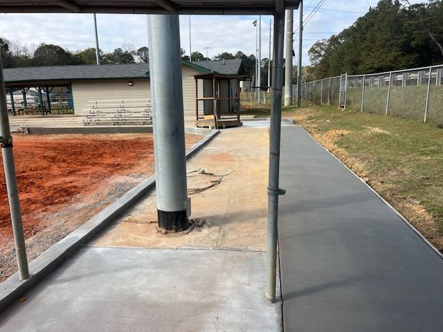 A freshly poured concrete sidewalk runs alongside a red dirt baseball field under a metal roof structure.