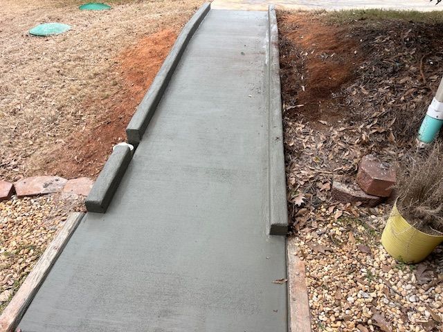 A newly poured gray concrete ramp with raised edges, surrounded by soil and gravel on an outdoor path.