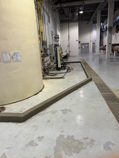 An industrial space showing a large tank with a concrete containment curb, piping, and a floor trench in a facility.