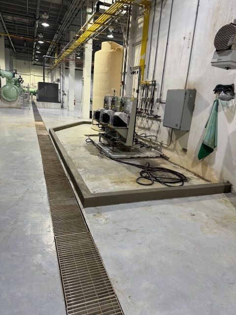 Industrial facility interior showing a metal floor trench drain, containment curbing, and pumps mounted on a concrete floor.
