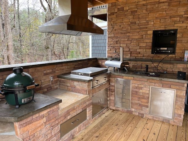 Outdoor kitchen on a wooden deck with a green kamado grill, stainless steel cooking appliances, and stone veneer walls.