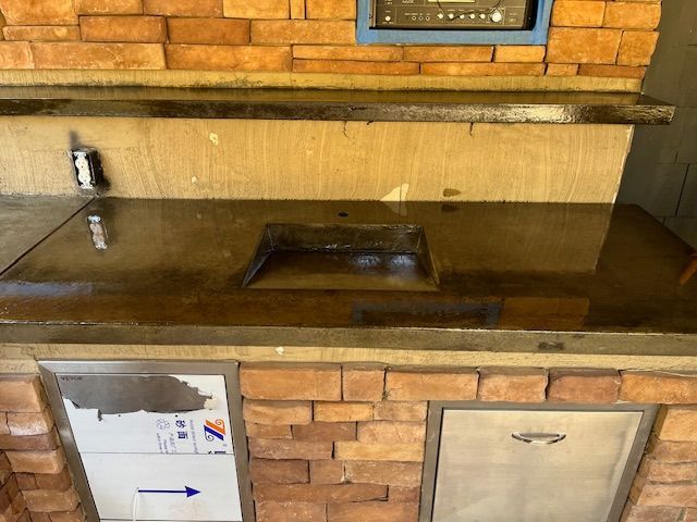 An outdoor kitchen counter with a dark concrete top, a square sink, stone brick facade, and stainless steel cabinetry.