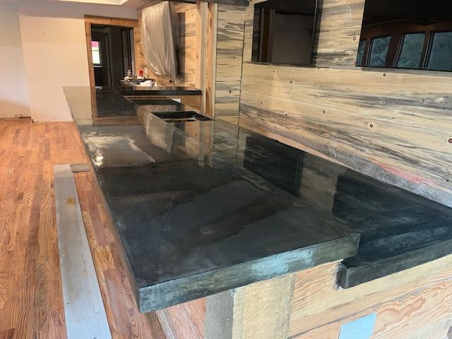 A dark, polished concrete countertop sits atop a rustic wooden base in a room with hardwood floors and wood paneling.