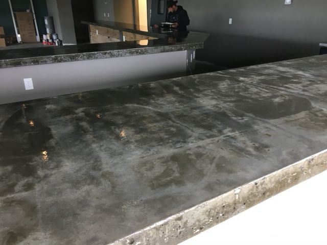 A close-up of a smooth, grey concrete bar top with a rough, unfinished front edge in a modern commercial space.