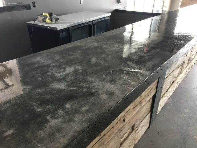 A polished dark gray concrete countertop with a rustic wood base and black metal corner trim in an interior space.